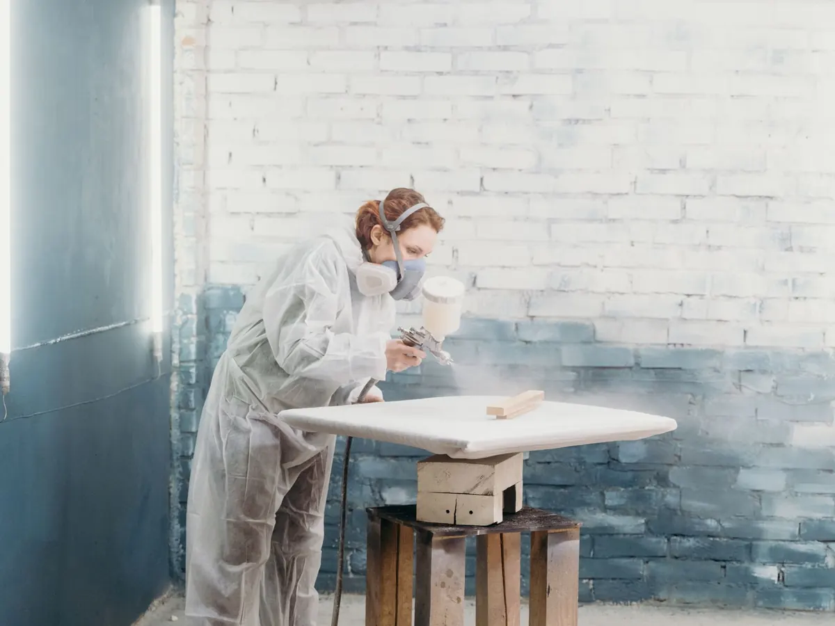 Decorator in a suit and respirator spraying a table top in a workshop
